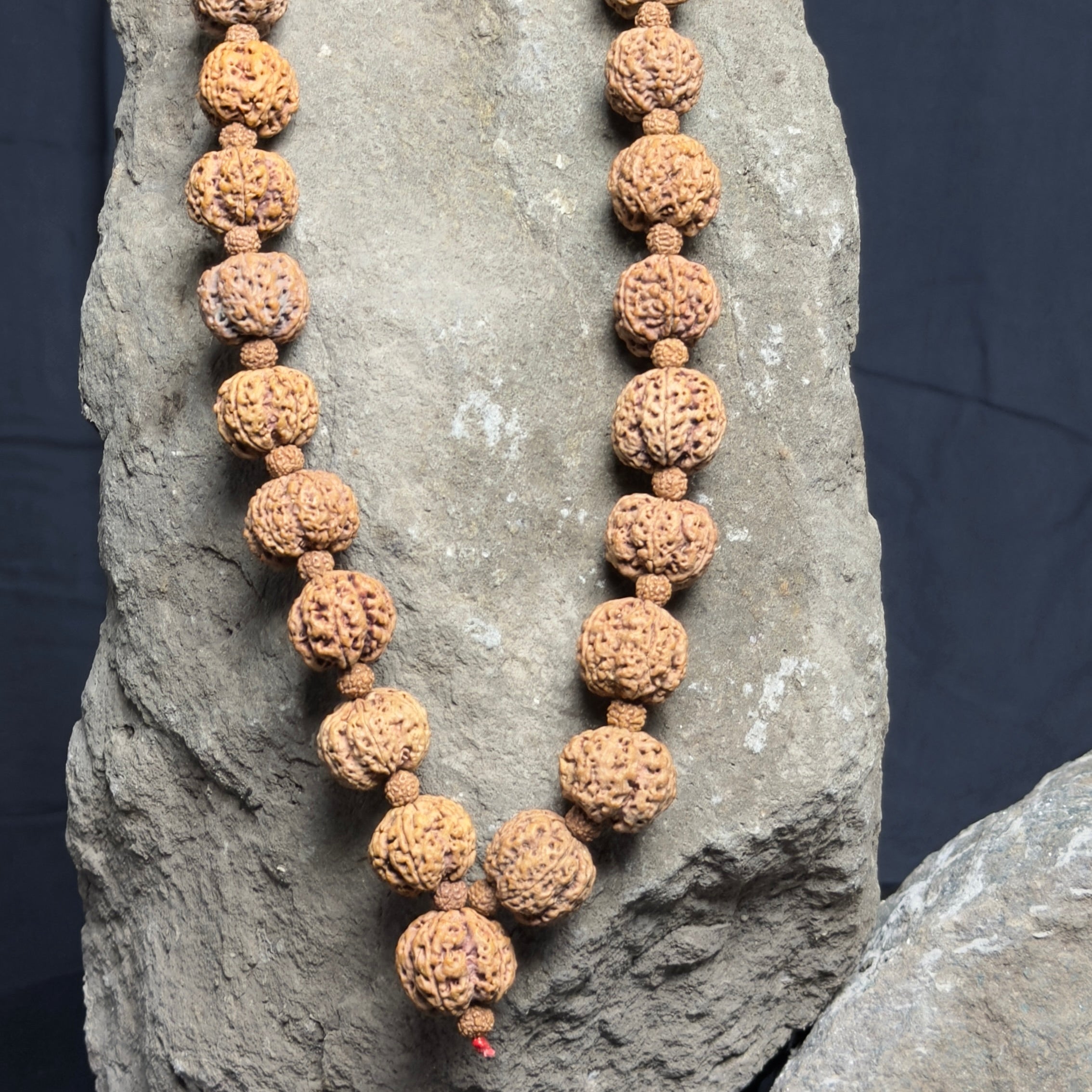 Natural brown Rudraksha beads mala with 5 Mukhi bead design, strung on tan thread, displayed on gray stone background.